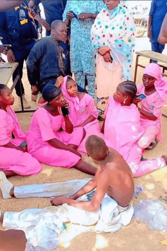 A group of people, mostly in pink attire, sit on the ground, engaging in conversation with others standing around them.