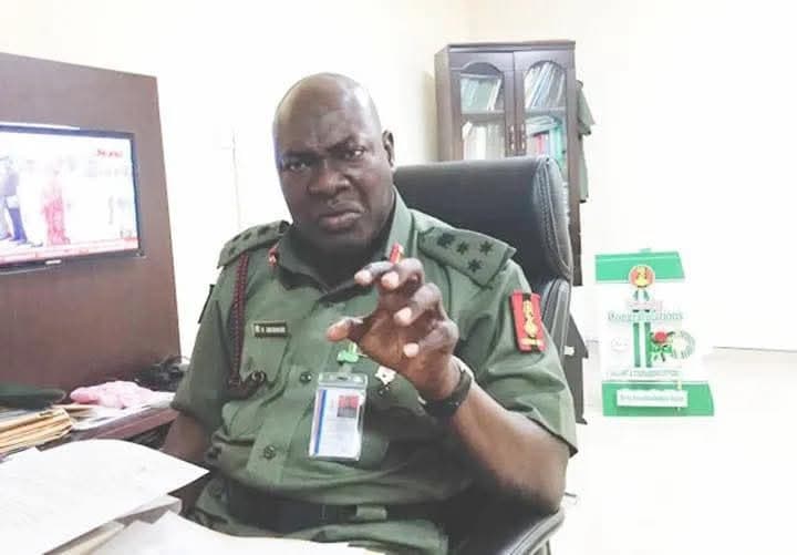 Man in military uniform gesturing while seated in an office with a TV in the background.