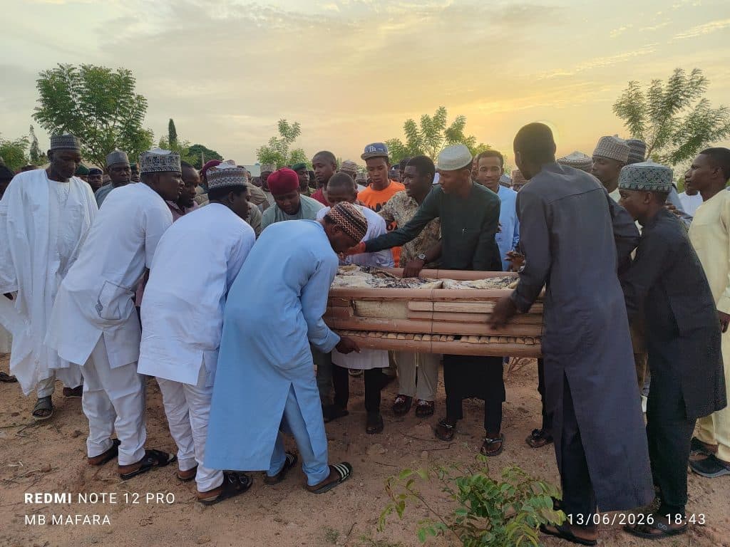 A group of people in traditional attire gather outdoors around a wrapped object at sunset.