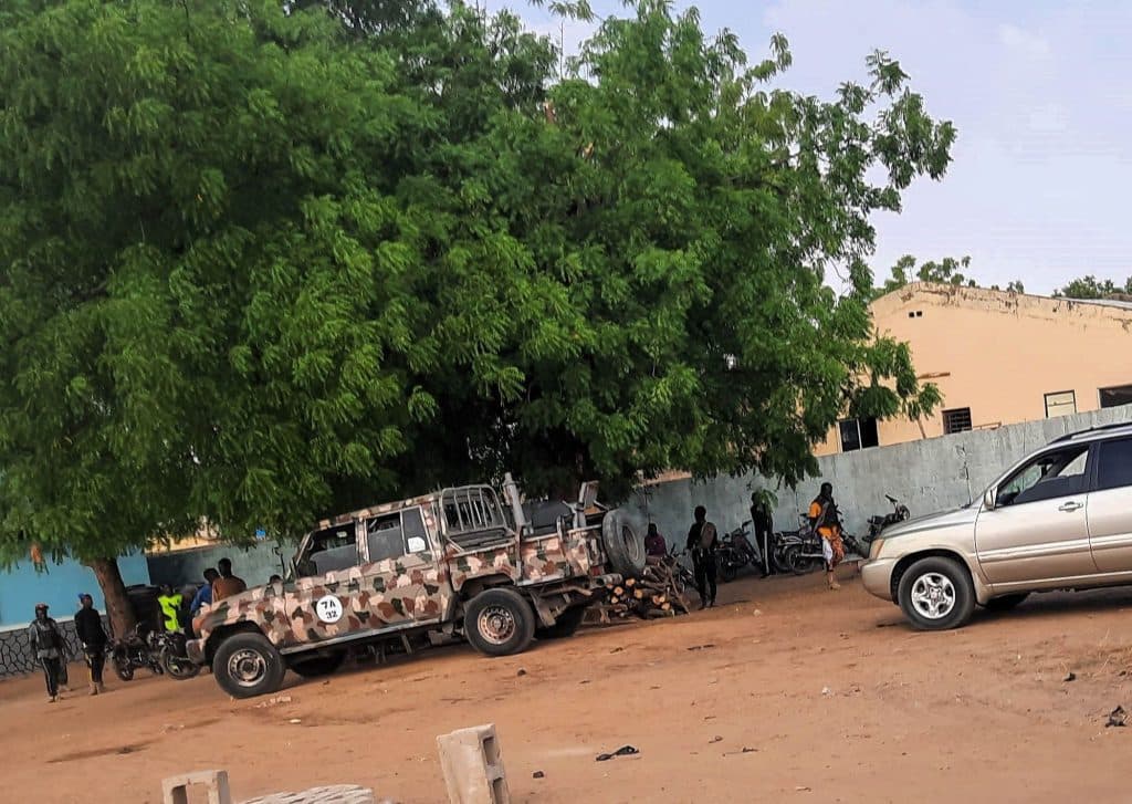 A camouflage-patterned military vehicle parked under a large tree, with people and motorcycles nearby. A beige SUV is also in the scene.