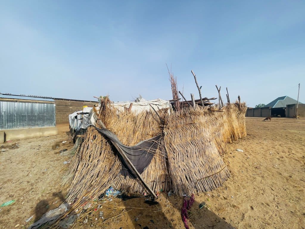 Tattered hut made of sticks and tarps in a dry, barren area, with other structures visible in the background under clear blue sky.