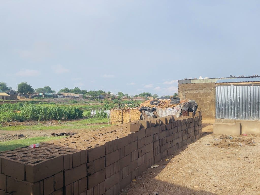 Open landscape with stacked bricks and metal sheds, green fields, and scattered rural structures under a clear sky.