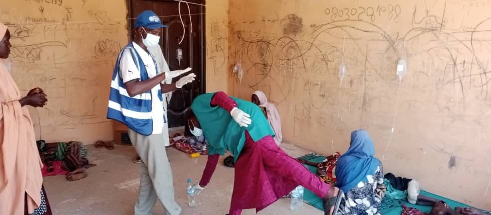 Healthcare workers assist individuals in a room with IV drips, graffiti on walls, and people sitting or lying on mats.