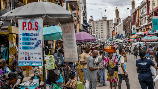 Busy street market scene with colorful umbrellas, a POS services sign, and people engaged in various activities.
