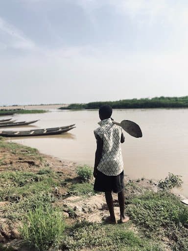 A person holding a paddle stands by a riverbank, looking at the water. Nearby, some wooden boats are docked.