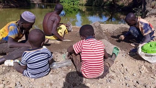Group of people by a pond, sorting through soil or sand, with bags and tools around them.