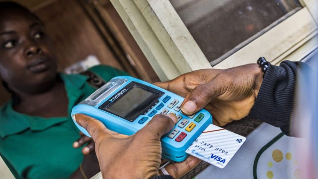 Person swiping a Visa card on a blue payment terminal at a counter, with another person observing.