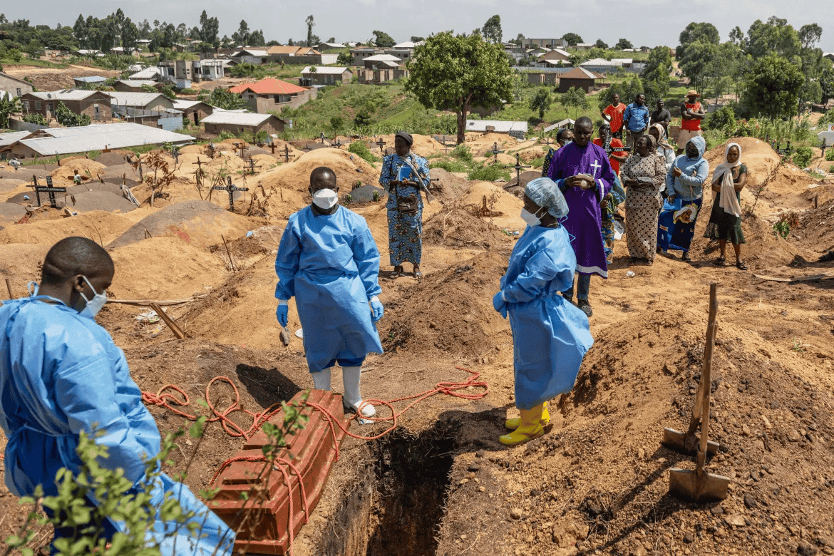 Healthcare workers in protective gear bury a coffin as onlookers stand by, in a hilly village cemetery under a blue sky.