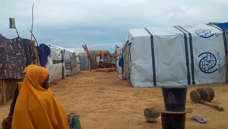 Person in yellow clothing sits near tents in a camp with cloudy skies above.