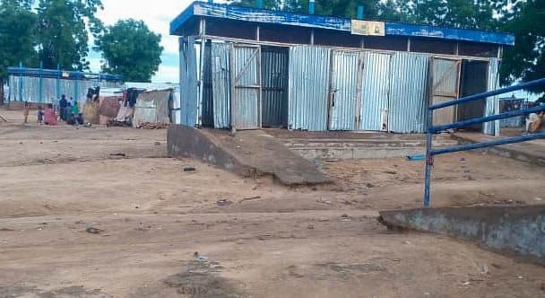 Corrugated metal building with ramps in a dirt lot; distant figures and trees in the background.