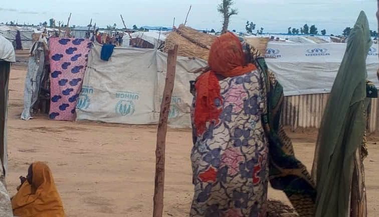 A woman in colorful clothing stands near makeshift shelters with UNHCR tarps in a dry area, another person sits nearby.