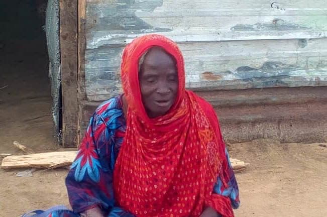 Elderly woman in a red headscarf and patterned dress sits in front of a weathered building.