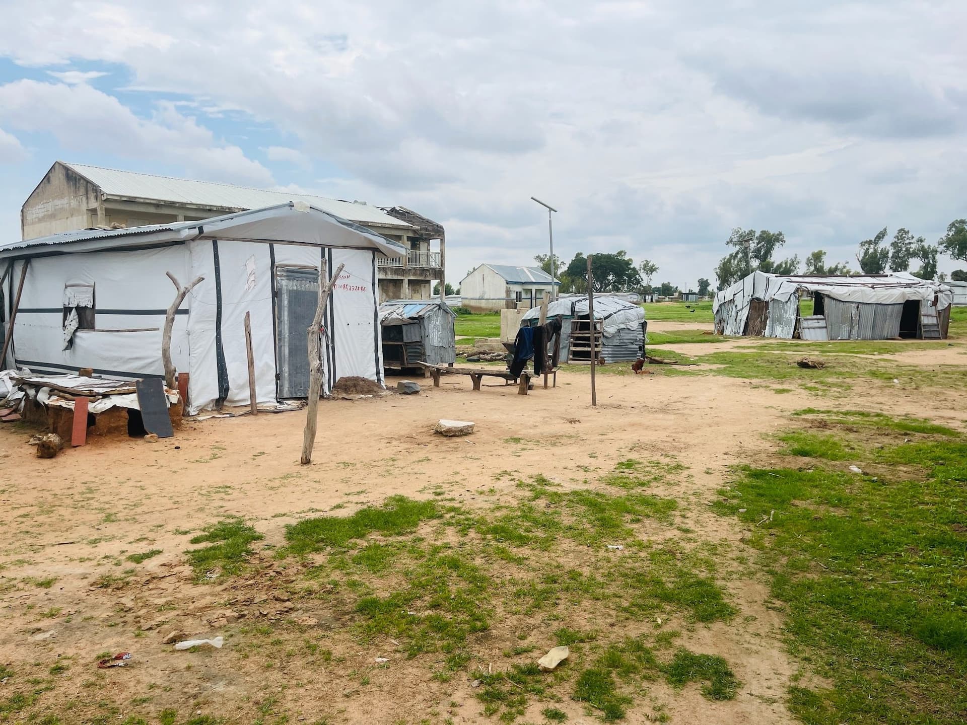 Simple structures with tin roofs and tarps on a grassy field under a cloudy sky.
