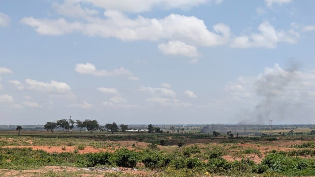 Expansive rural landscape with a clear sky, scattered clouds, distant trees, and a plume of smoke rising on the right.