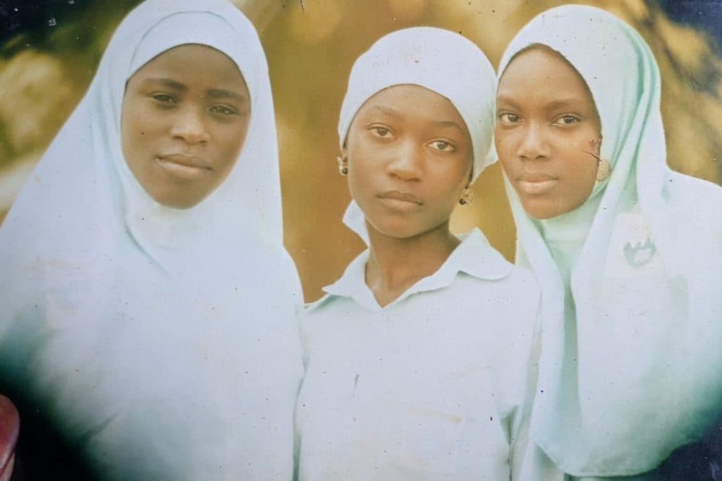Three young girls in light-colored hijabs and uniforms stand close together, looking at the camera.