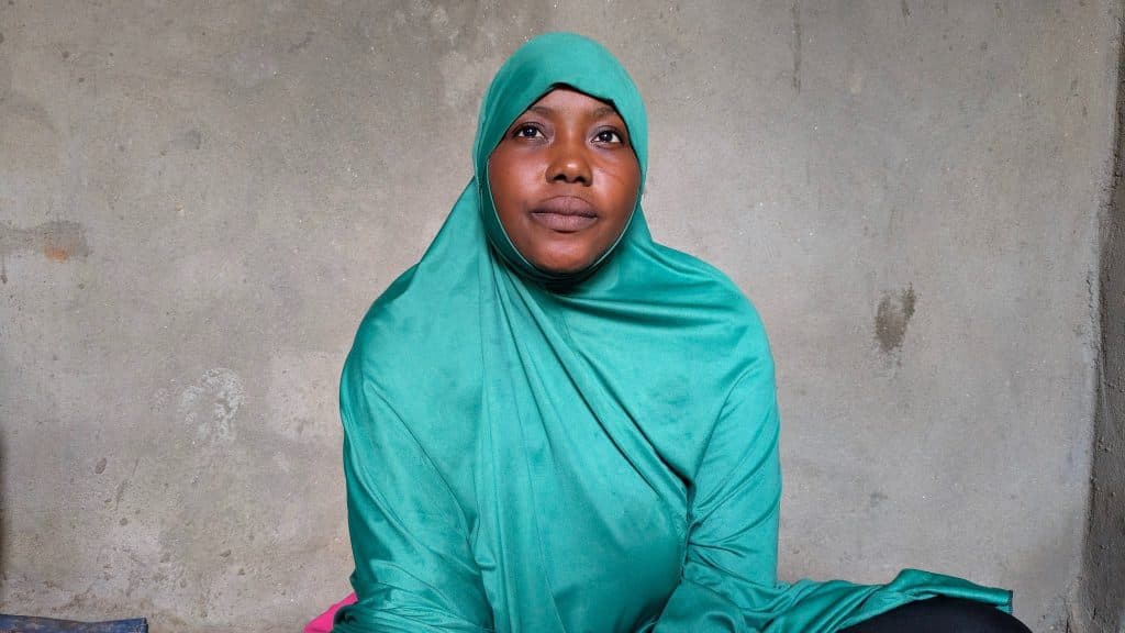 Person wearing a green hijab sitting against a plain gray wall.