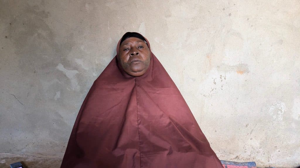 A person wearing a brown cloak sits against a plain wall.