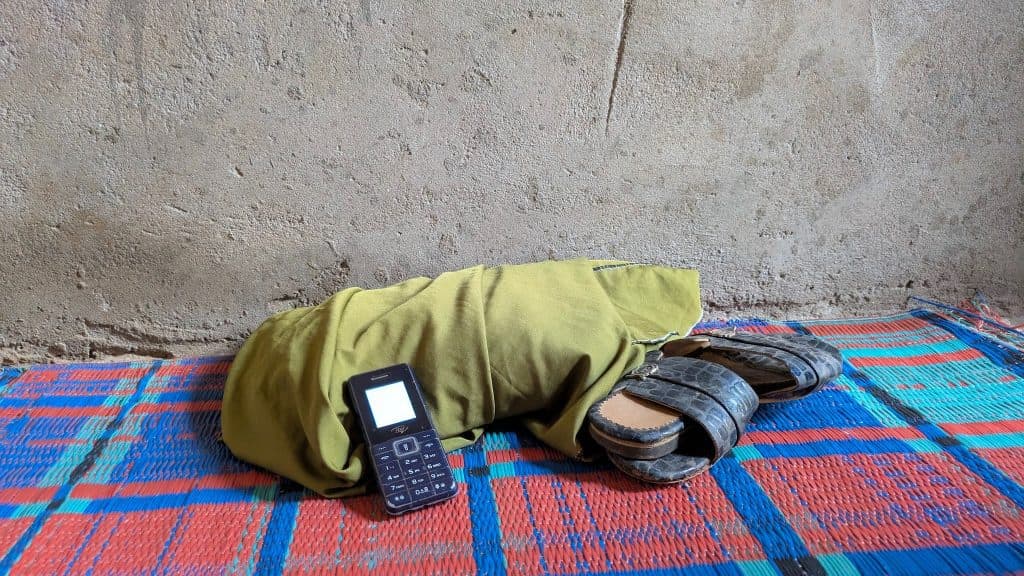 Cell phone, green cloth, and sandals on a colorful mat against a bare wall.