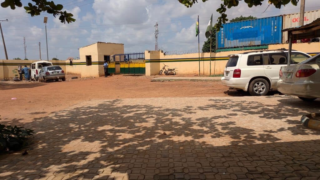Parking area with cars and a bicycle by a gated building. Flags are flying, and people are gathered near a wall.