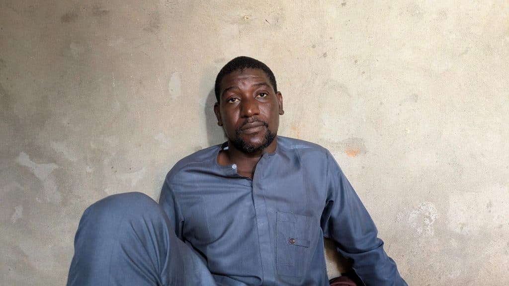 Man in gray shirt sitting against a textured wall, looking directly at the camera.