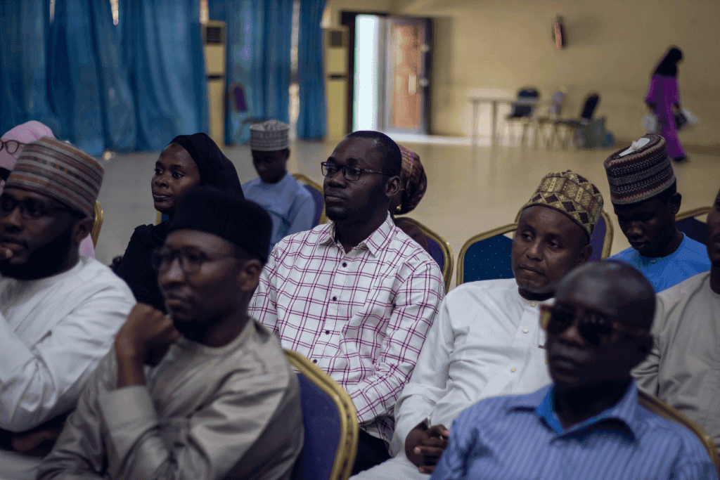 People seated attentively in a room with blue curtains and empty chairs in the background.