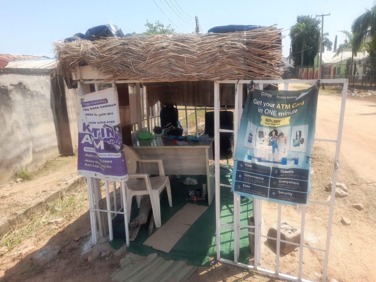 Small outdoor booth with straw roof, advertising ATM and banking services. Two banners hang at the entrance, with chairs and a table inside.