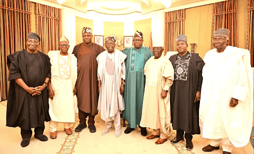 A group of eight men wearing traditional Nigerian attire stand together, posing for a photo in an elegantly decorated room.