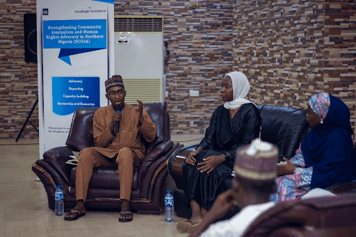 People in discussion at a community journalism and human rights event, sitting on chairs with a banner in the background.