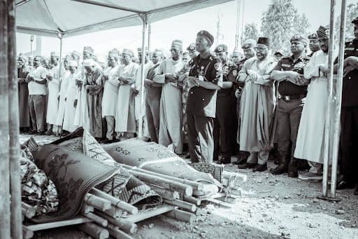 A group of people, some in uniform, stand in prayer under a tent beside two covered stretchers outdoors.