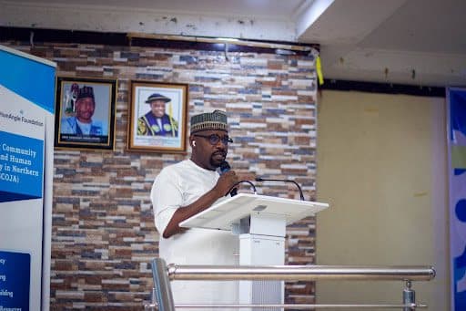 A person giving a speech at a podium indoors, with framed portraits and a banner in the background.