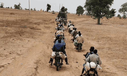 A group of people on motorcycles riding on a dirt path through a dry rural landscape with trees in the distance.