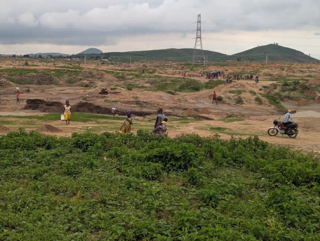 People and motorbikes on a dirt path in a rural landscape, with green plants and hills in the background under a cloudy sky.