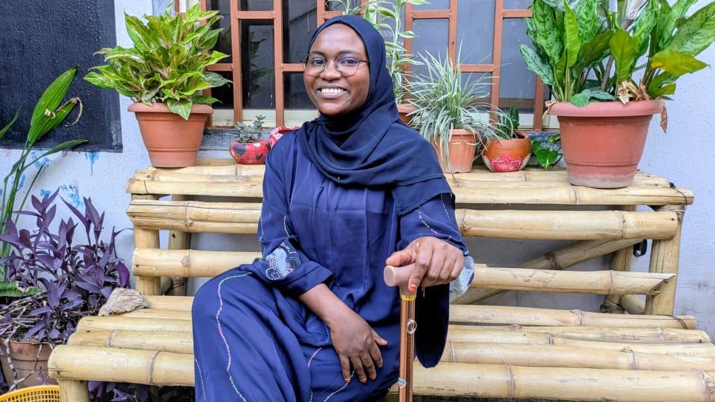 A woman in a hijab smiles while sitting on a bamboo bench surrounded by plants.