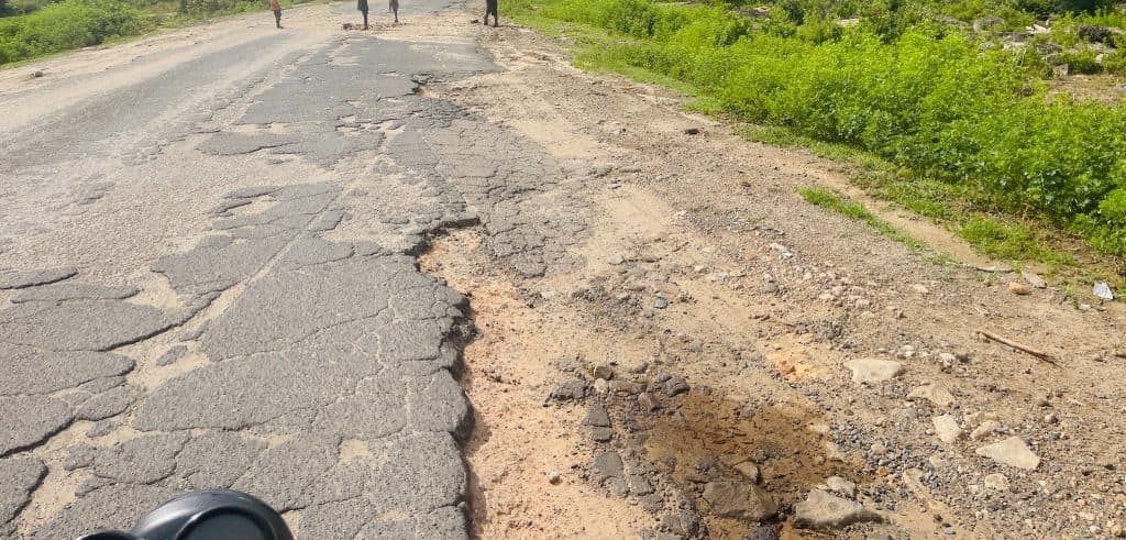 Cracked, uneven road with patches, surrounded by green vegetation. Three people walking in the distance.