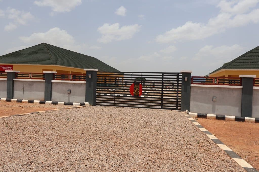School entrance with a metal gate, paved driveway, and buildings with green roofs under a partly cloudy sky.