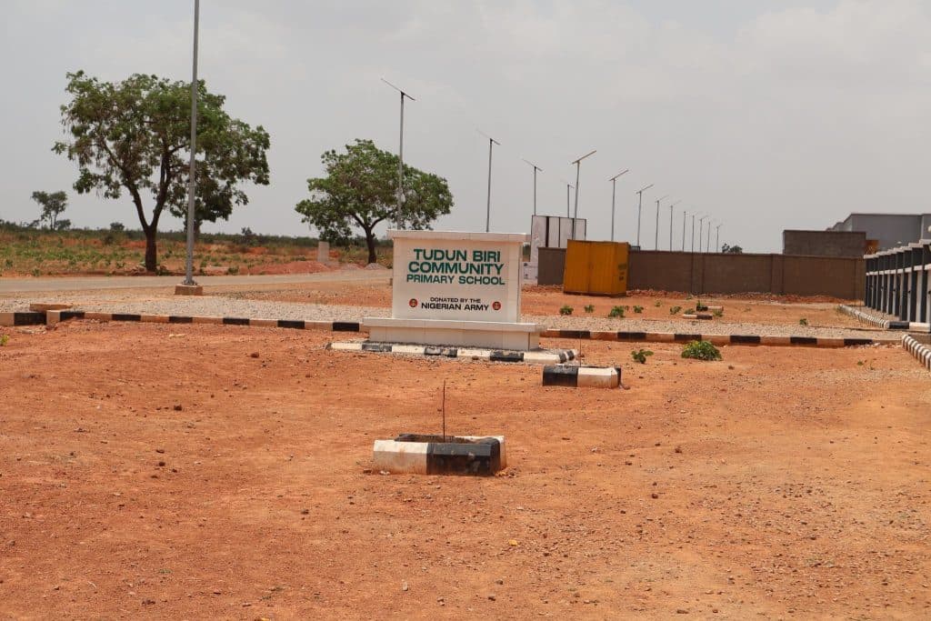 Sign for Tudun Biri Community Primary School, donated by the Nigerian Army, on a dirt path with trees and distant buildings.