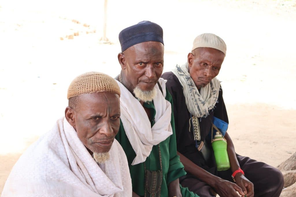 Three elderly men sit together, wearing traditional clothing and hats, with neutral expressions in an outdoor setting.