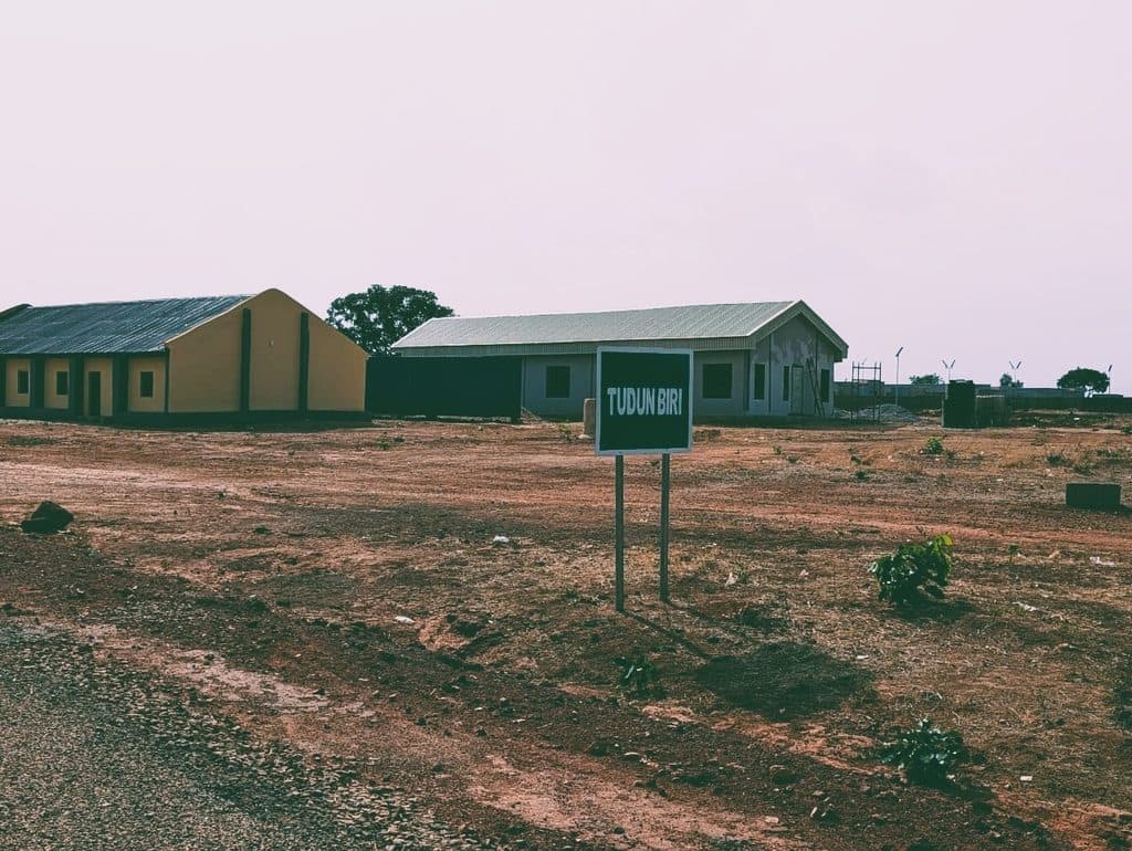 A rural area with two buildings, a sign saying "Tudun Biri," dry ground, and sparse vegetation under a clear sky.
