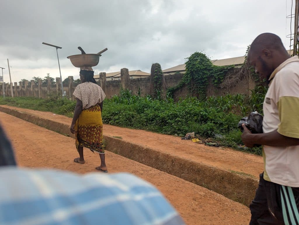 Person walking on dirt road, carrying a large metal pan on their head. Another person is on the right holding a plastic bag.