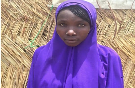 A girl in a purple hijab is standing in front of a straw hut.