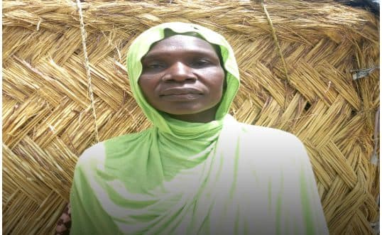 A woman in a green shawl standing in front of a straw hut.