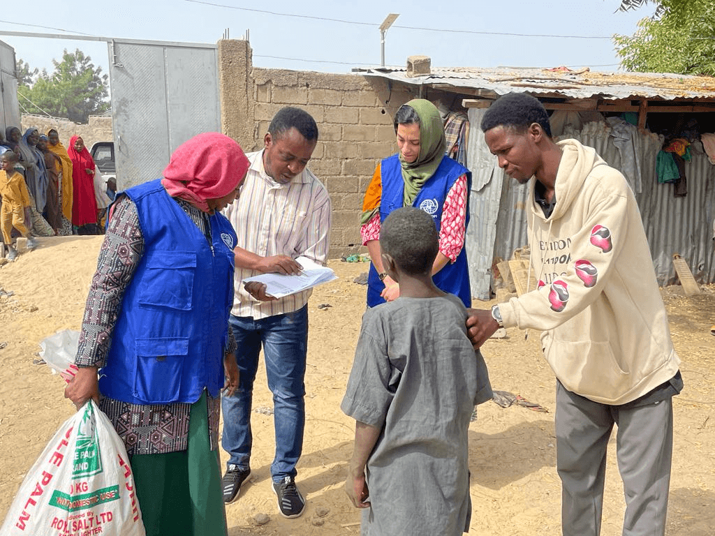 Aid workers engage with locals in a dusty village, carrying supplies and documents.