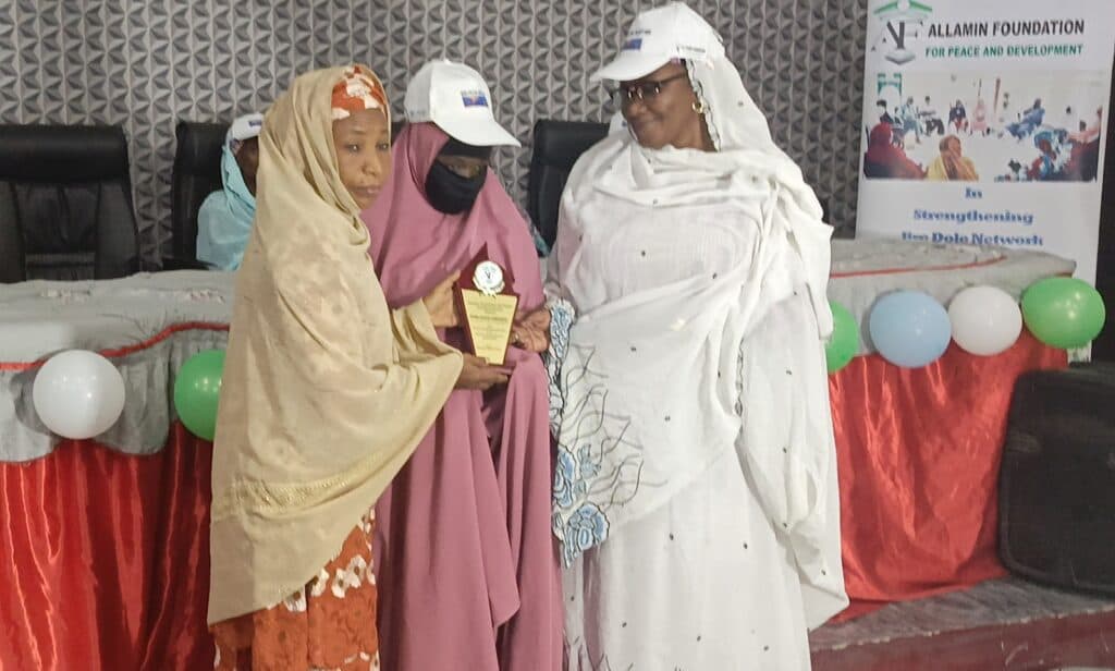 Three women at an event, one receiving an award, with balloons and a banner in the background.
