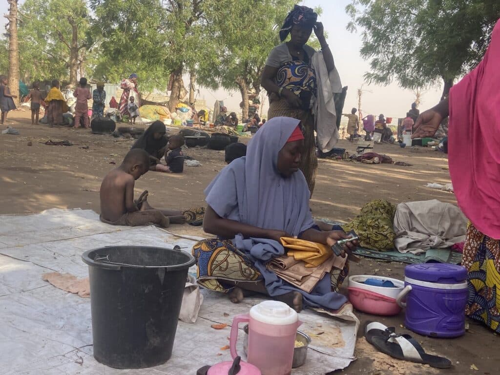 Outdoor setting with people seated on the ground, some with cooking pots, in a daily life scene in a rural environment.