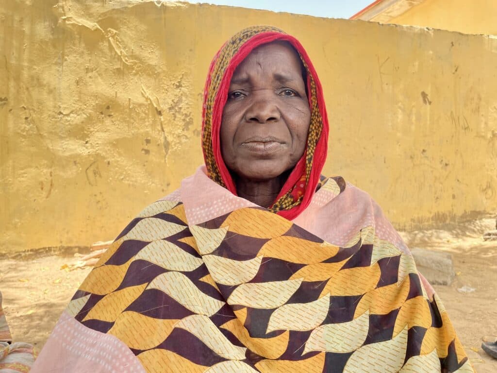 Elderly woman with a headscarf wrapped around her, wearing a patterned shawl, in front of a yellow wall.