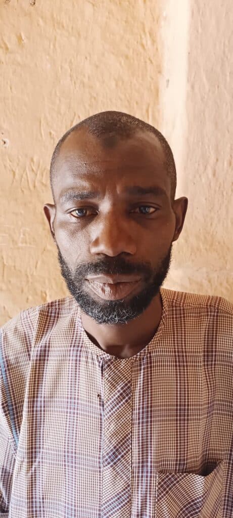 Man with a beard in a plaid shirt standing against a textured wall.