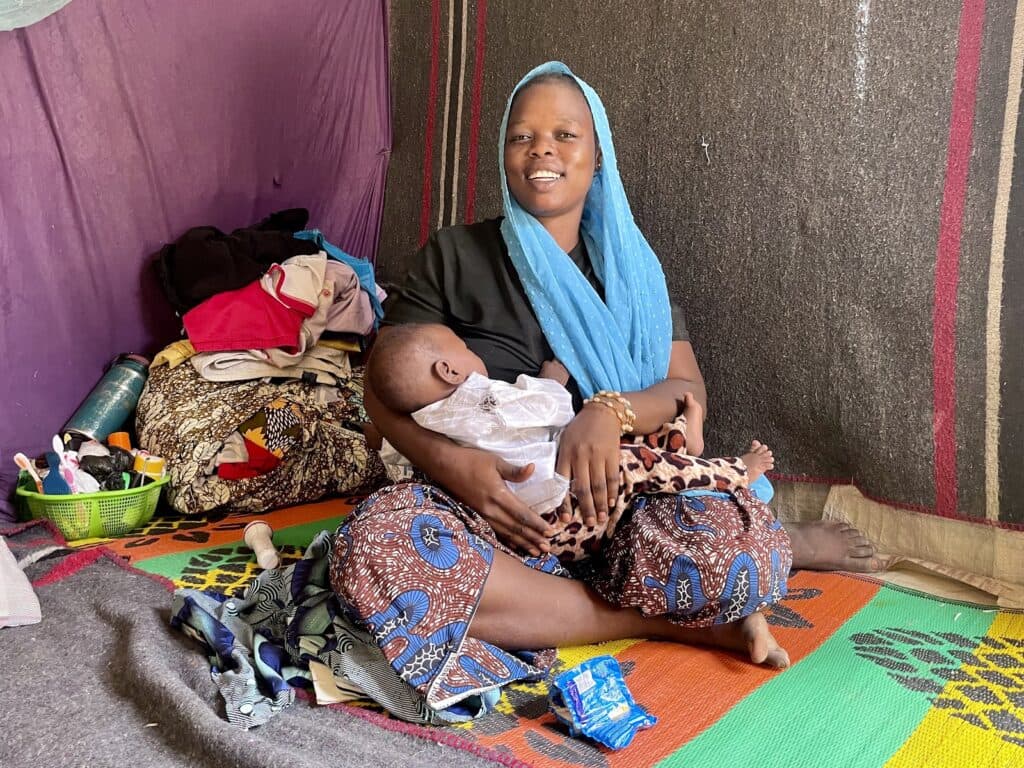 Mother in colorful attire holding a baby indoors, sitting on a woven mat, with household items nearby.