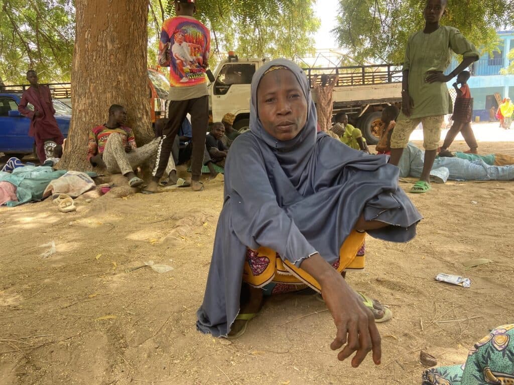 A woman in traditional clothing sitting on the ground under a tree with other people in a relaxed outdoor setting.