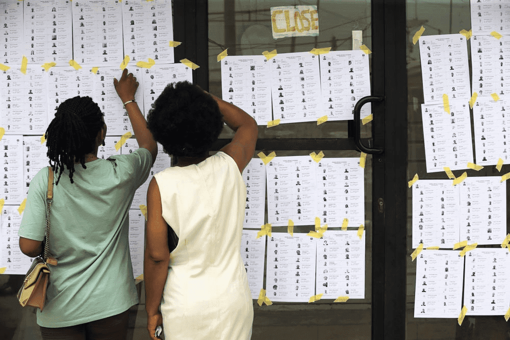 Two people checking a wall of posted papers outside a closed door.