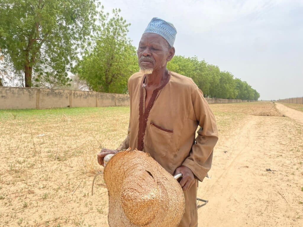 Elderly man in traditional attire holding a straw hat, standing on a dusty path with trees in the background.
